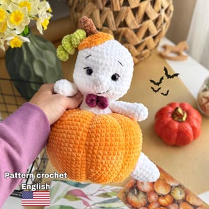 Può includere: Un peluche a forma di fantasma lavorato all'uncinetto bianco con occhi neri e un piccolo papillon viola, seduto sopra una grande zucca lavorata all'uncinetto arancione. Il fantasma ha un gambo marrone e viticci verdi sulla testa. Il testo Pattern crochet English è visibile.