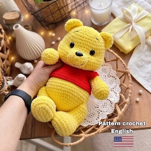 May include: A handmade yellow crochet bear wearing a red shirt. The bear has black eyes and a black nose. The image includes the text "Pattern crochet English" and an American flag.