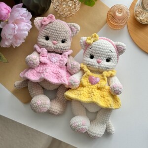 Pode incluir: Duas bonecas de gato de croch&ecirc; feitas &agrave; m&atilde;o. Um gato &eacute; bege com um vestido e la&ccedil;o rosa, o outro &eacute; branco com um vestido e la&ccedil;o amarelo. Ambas as bonecas t&ecirc;m enfeites em forma de cora&ccedil;&atilde;o em seus vestidos e detalhes rosa nas patas.