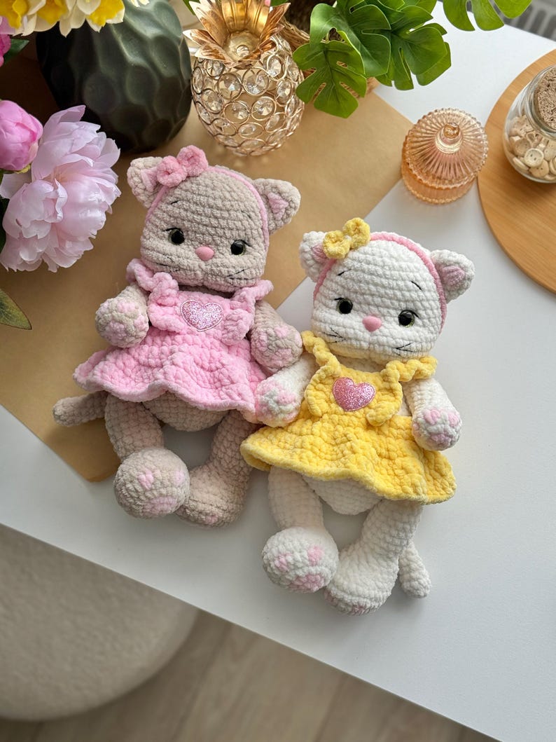 Pode incluir: Duas bonecas de gato de croch&ecirc; feitas &agrave; m&atilde;o. Um gato &eacute; bege com um vestido e la&ccedil;o rosa, o outro &eacute; branco com um vestido e la&ccedil;o amarelo. Ambas as bonecas t&ecirc;m enfeites em forma de cora&ccedil;&atilde;o em seus vestidos e detalhes rosa nas patas.