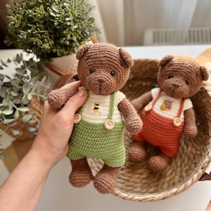 May include: Two crocheted teddy bears. One bear is held in a hand and wears green overalls with a white shirt featuring a bee graphic. The other bear is in a woven basket and wears orange overalls. Both bears are brown.