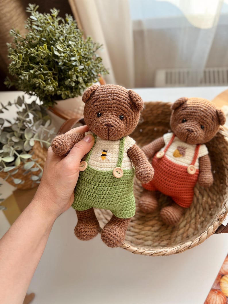 May include: Two crocheted teddy bears. One bear is held in a hand and wears green overalls with a white shirt featuring a bee graphic. The other bear is in a woven basket and wears orange overalls. Both bears are brown.