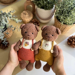 May include: Two handmade crochet teddy bears. The bears are brown with white shirts and overalls. One bear has rust-colored overalls, and the other has yellow overalls. Each bear has a small floral design on the shirt.
