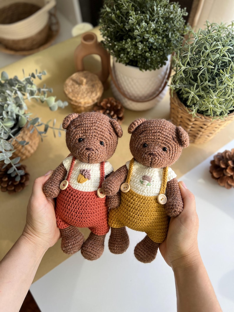 May include: Two handmade crochet teddy bears. The bears are brown with white shirts and overalls. One bear has rust-colored overalls, and the other has yellow overalls. Each bear has a small floral design on the shirt.