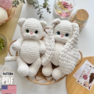May include: Two crocheted stuffed animals, a white cat and a white bunny, are sitting in a wicker basket. Both animals have large, round eyes and are made with a soft, fluffy yarn.