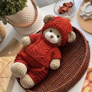 May include: A handmade crochet teddy bear wearing a red hooded sweater and matching pants. The bear has tan-colored paws, ears, and snout. It is lying in a brown woven basket. The bear's outfit has a cable knit design.