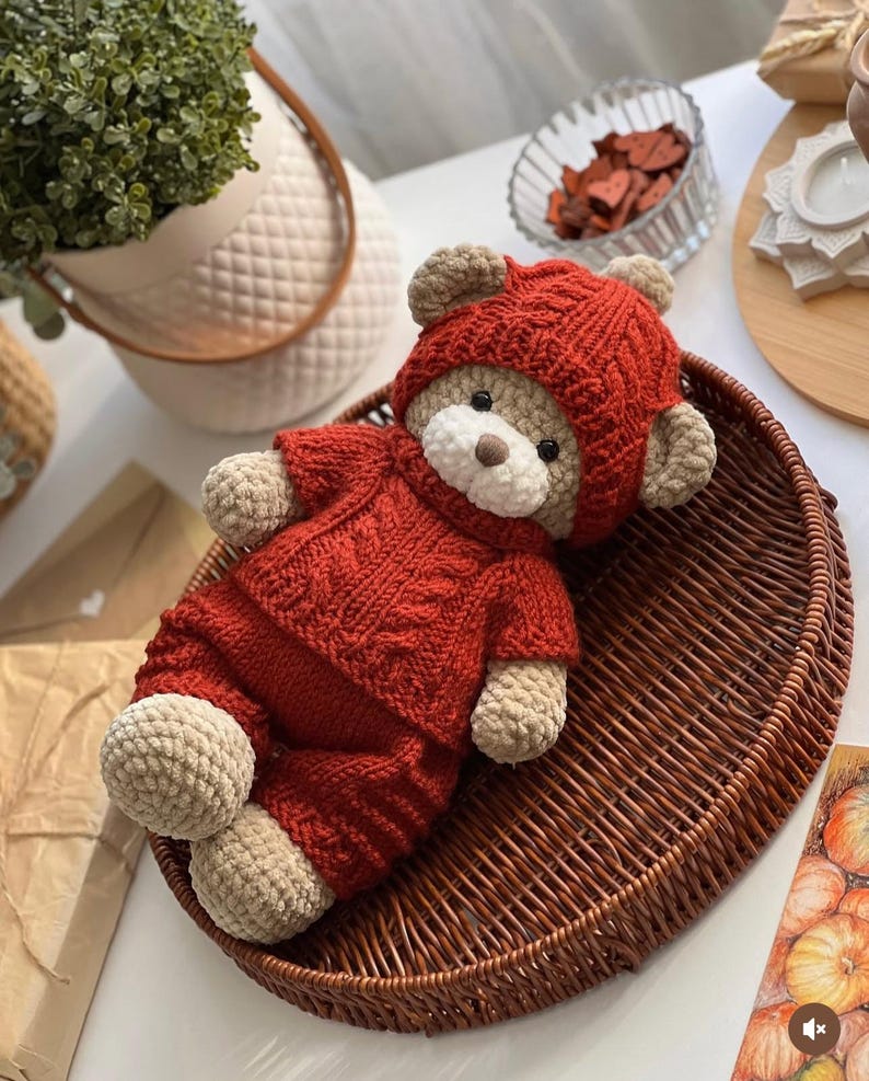 May include: A handmade crochet teddy bear wearing a red hooded sweater and matching pants. The bear has tan-colored paws, ears, and snout. It is lying in a brown woven basket. The bear's outfit has a cable knit design.