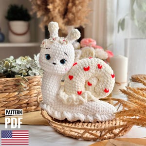 以下が含まれることがあります： 手作りの白いかぎ針編みのカタツムリ。フリルエッジと赤いハートのアクセントが特徴です。カタツムリは青い目を持ち、花の冠をかぶっています。画像には「PATTERN PDF」の文字とアメリカ国旗が含まれています。