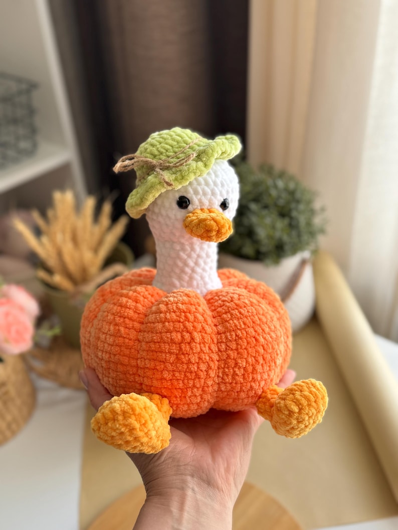 May include: A handmade plush goose with a white head, orange beak, and a green hat. The goose's body is orange and shaped like a pumpkin. The plush toy is held in a person's hand.