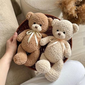 May include: Two crocheted teddy bears, one brown and one cream, sitting in a wicker basket. Both bears have a ribbon tied around their necks.