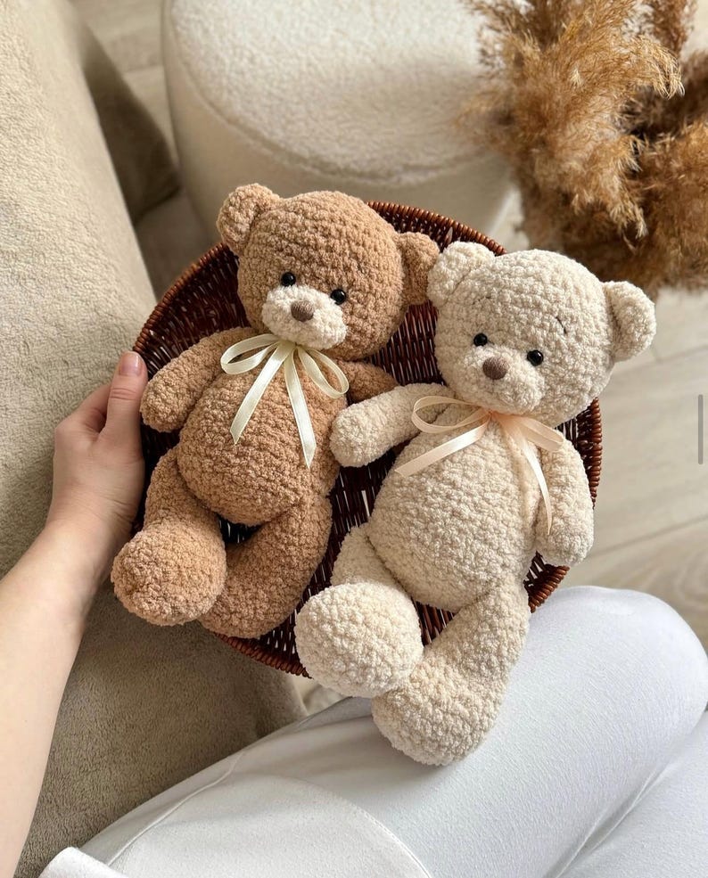 May include: Two crocheted teddy bears, one brown and one cream, sitting in a wicker basket. Both bears have a ribbon tied around their necks.