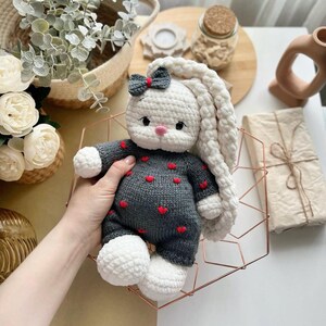 Puede incluir: Mu&ntilde;eca de conejo de ganchillo hecha a mano con cabeza y orejas largas blancas, cuerpo gris adornado con corazones rojos y pies blancos. El conejito tiene un peque&ntilde;o lazo en la cabeza y se sostiene en una cesta de alambre geom&eacute;trica.
