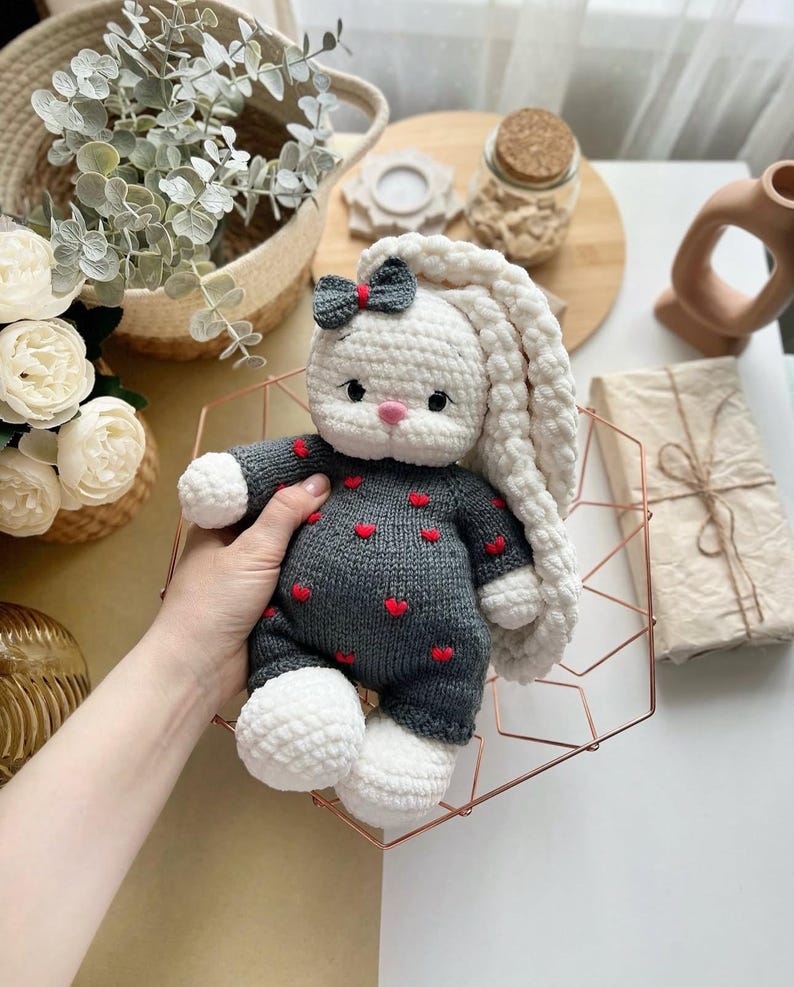 Puede incluir: Mu&ntilde;eca de conejo de ganchillo hecha a mano con cabeza y orejas largas blancas, cuerpo gris adornado con corazones rojos y pies blancos. El conejito tiene un peque&ntilde;o lazo en la cabeza y se sostiene en una cesta de alambre geom&eacute;trica.