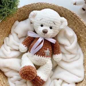May include: A white crocheted teddy bear with brown accents and a silver star on its chest. The bear is wearing a white ribbon around its neck and is sitting in a white blanket.