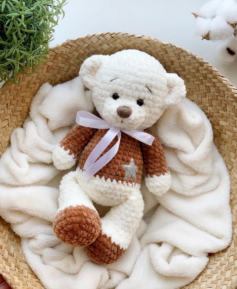 May include: A white crocheted teddy bear with brown accents and a silver star on its chest. The bear is wearing a white ribbon around its neck and is sitting in a white blanket.