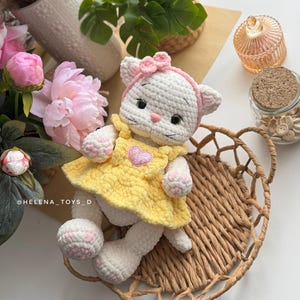 Pode incluir: Boneca de gato de croch&ecirc; branco feita &agrave; m&atilde;o, vestindo um vestido amarelo com um aplique de cora&ccedil;&atilde;o rosa. O gato tem uma faixa de la&ccedil;o rosa e est&aacute; sentado em uma cesta de vime. As patas e orelhas da boneca t&ecirc;m detalhes em rosa. &Eacute; um brinquedo macio.