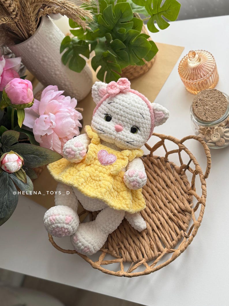 Pode incluir: Boneca de gato de croch&ecirc; branco feita &agrave; m&atilde;o, vestindo um vestido amarelo com um aplique de cora&ccedil;&atilde;o rosa. O gato tem uma faixa de la&ccedil;o rosa e est&aacute; sentado em uma cesta de vime. As patas e orelhas da boneca t&ecirc;m detalhes em rosa. &Eacute; um brinquedo macio.