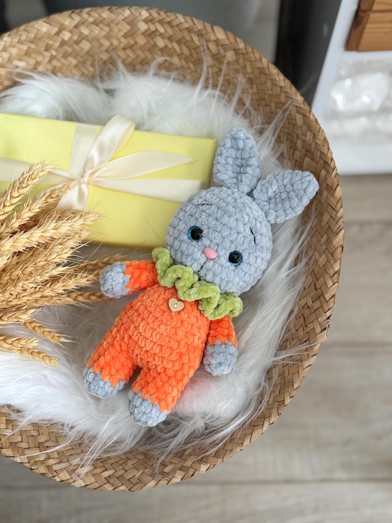May include: A gray crocheted bunny wearing an orange crocheted outfit with a green and yellow crocheted collar. The bunny is sitting in a woven basket.