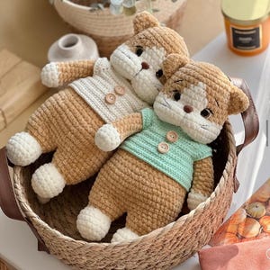 Pu&ograve; includere: Tre bambole a forma di gatto all'uncinetto fatte a mano in un cestino di vimini. Le bambole hanno corpi color beige, dettagli bianchi e indossano maglioni color crema e verde menta. Le bambole hanno chiusure con bottoni e sono in un cestino intrecciato.