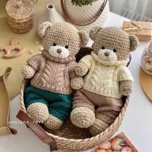 May include: Two handmade crochet teddy bears in a woven basket. One bear wears a pink sweater and teal pants, the other a cream sweater and brown pants. Both have tan heads and white muzzles. The bears are a charming addition to any nursery or child's room.