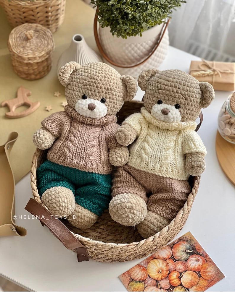 May include: Two handmade crochet teddy bears in a woven basket. One bear wears a pink sweater and teal pants, the other a cream sweater and brown pants. Both have tan heads and white muzzles. The bears are a charming addition to any nursery or child's room.