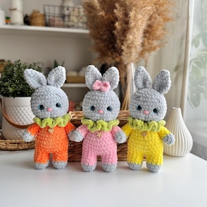 May include: Three crocheted gray bunny toys wearing different colored outfits. The bunny on the left is wearing orange, the middle bunny is wearing pink, and the bunny on the right is wearing yellow.