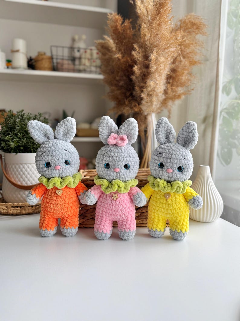 May include: Three crocheted gray bunny toys wearing different colored outfits. The bunny on the left is wearing orange, the middle bunny is wearing pink, and the bunny on the right is wearing yellow.