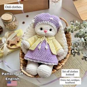 May include: A handmade crochet bunny doll wearing a purple dress, yellow cardigan, and a matching purple hat. The bunny has long white ears and is seated in a woven basket. The text "Onli clothes" is visible.