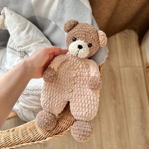May include: A crocheted brown and pink teddy bear wearing a pink crocheted outfit with brown buttons. The bear is sitting in a wicker basket.