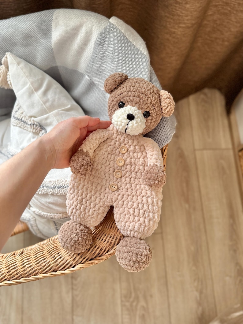 May include: A crocheted brown and pink teddy bear wearing a pink crocheted outfit with brown buttons. The bear is sitting in a wicker basket.