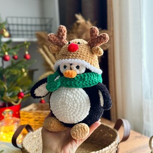 May include: A handmade crochet penguin wearing a reindeer hat and a green scarf. The penguin has a white belly, black wings, and tan feet. The reindeer hat is tan with brown antlers and a red nose. The penguin is a festive holiday decoration.