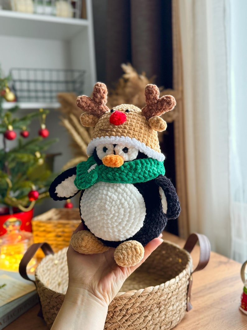 May include: A handmade crochet penguin wearing a reindeer hat and a green scarf. The penguin has a white belly, black wings, and tan feet. The reindeer hat is tan with brown antlers and a red nose. The penguin is a festive holiday decoration.
