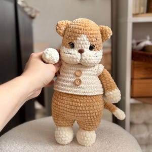 Pu&ograve; includere: Bambola a forma di gatto all'uncinetto fatta a mano, nei toni del marrone e del crema. Il gatto indossa un gilet color crema con bottoni in legno. Il gatto ha muso, zampe e punta della coda bianchi. La bambola &egrave; su una superficie bianca strutturata.