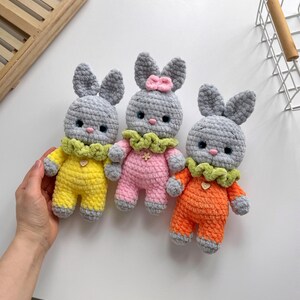 May include: Three crocheted bunny plushies with gray fur, each wearing a different colored outfit: yellow, pink, and orange. The bunnies have green collars and small hearts on their chests.