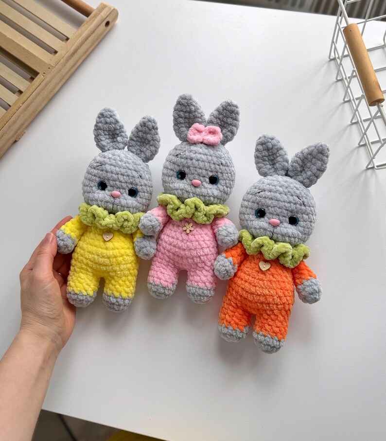 May include: Three crocheted bunny plushies with gray fur, each wearing a different colored outfit: yellow, pink, and orange. The bunnies have green collars and small hearts on their chests.