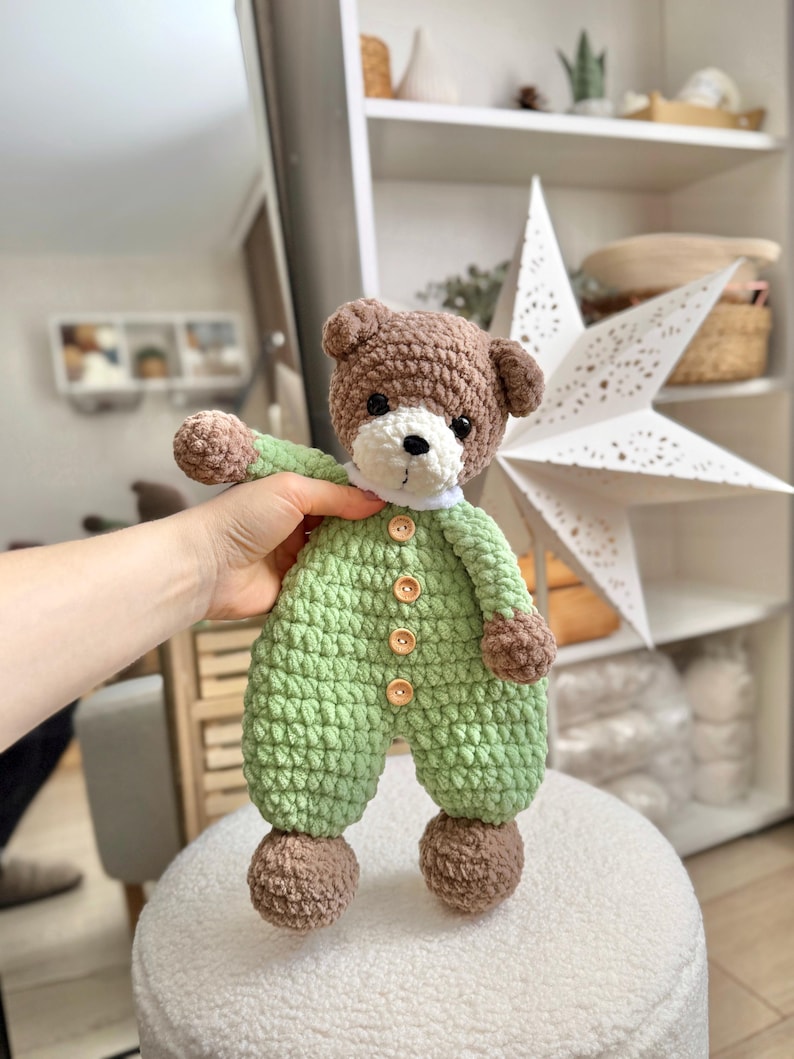 May include: A crocheted brown bear wearing a green jumpsuit with brown buttons. The bear has a white face with black eyes and a black nose.