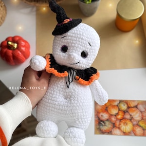 May include: A handmade white crochet ghost doll wearing a black witch's hat and a black and orange collar. The doll has purple eyes and a smiling face. The doll is a Halloween-themed item.