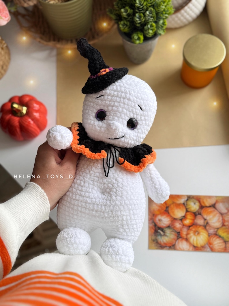 May include: A handmade white crochet ghost doll wearing a black witch's hat and a black and orange collar. The doll has purple eyes and a smiling face. The doll is a Halloween-themed item.