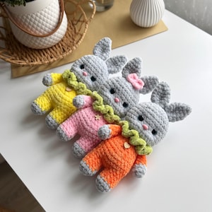 May include: Three crocheted gray bunny plushies wearing different colored outfits. The bunnies are wearing yellow, pink, and orange outfits with green collars and decorative accents.