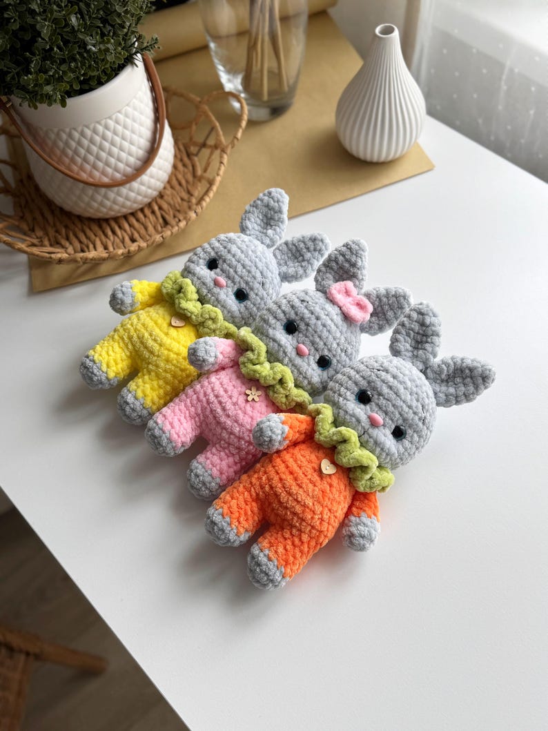 May include: Three crocheted gray bunny plushies wearing different colored outfits. The bunnies are wearing yellow, pink, and orange outfits with green collars and decorative accents.