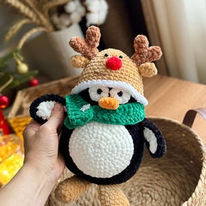 May include: A handmade crochet penguin wearing a reindeer hat and green scarf. The penguin has a white belly, black body, and tan feet. The reindeer hat is tan with brown antlers and a red nose. The scarf has a white snowflake.