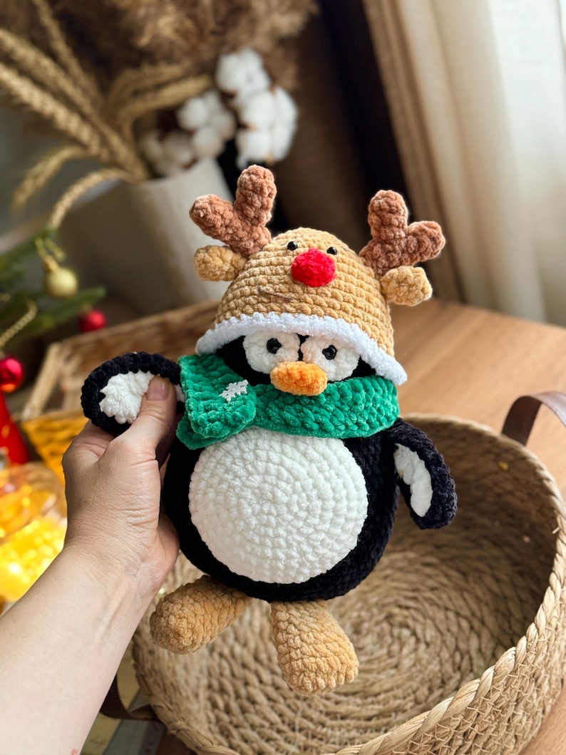 May include: A handmade crochet penguin wearing a reindeer hat and green scarf. The penguin has a white belly, black body, and tan feet. The reindeer hat is tan with brown antlers and a red nose. The scarf has a white snowflake.