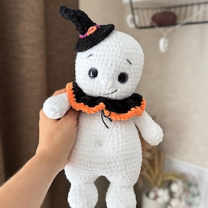 May include: A handmade white crochet ghost doll wearing a black witch's hat with orange and purple details. The ghost has a black collar with orange trim and black eyes. A Halloween-themed item.