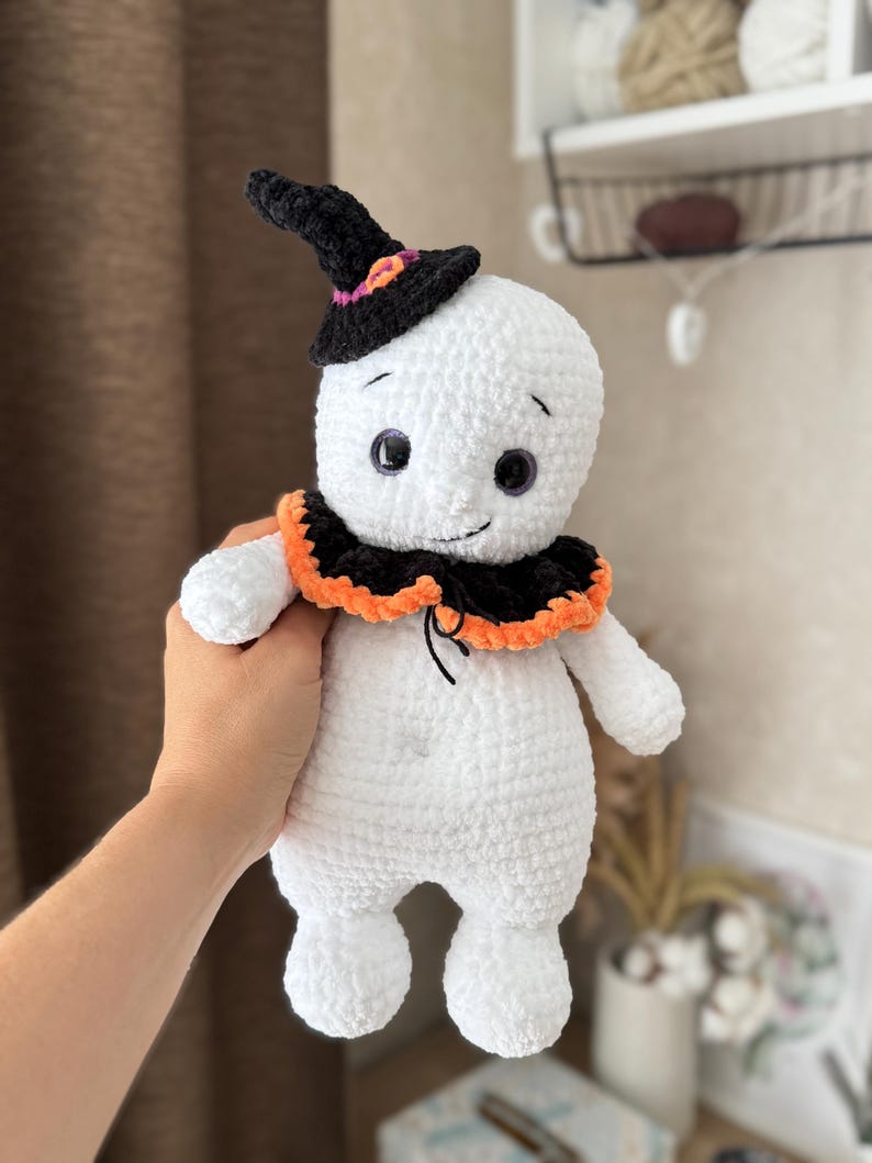 May include: A handmade white crochet ghost doll wearing a black witch's hat with orange and purple details. The ghost has a black collar with orange trim and black eyes. A Halloween-themed item.