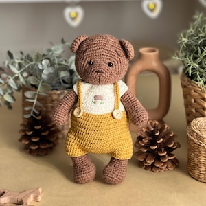 May include: A handmade crochet teddy bear with brown fur, wearing a white shirt with a floral design and mustard yellow overalls. The bear has button details and is standing on a brown surface. The background includes decorative items.