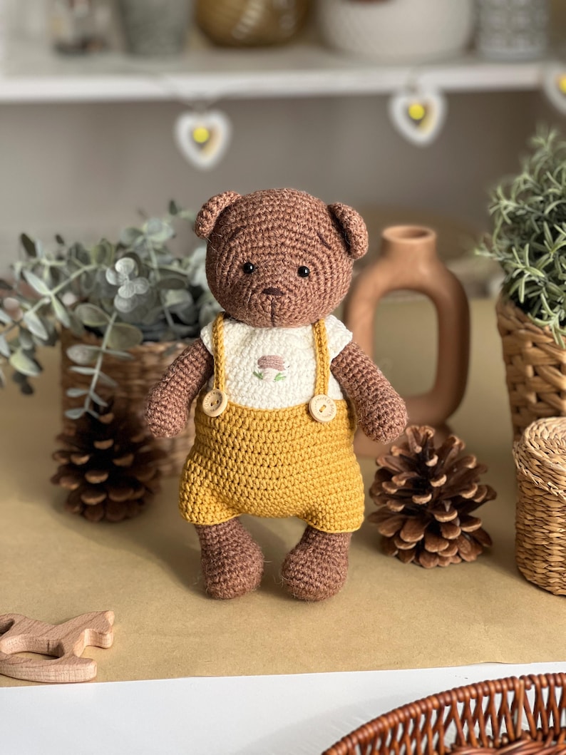 May include: A handmade crochet teddy bear with brown fur, wearing a white shirt with a floral design and mustard yellow overalls. The bear has button details and is standing on a brown surface. The background includes decorative items.