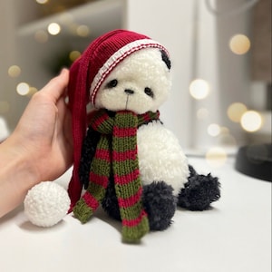 May include: A white and black panda bear plush toy wearing a red and white knit hat and a green and red striped scarf. The toy is sitting on a white surface.