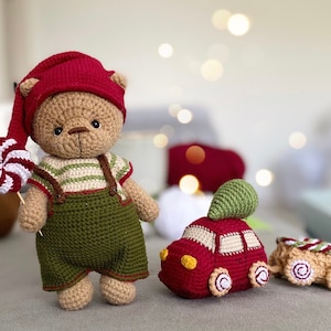 Puede incluir: Un oso de peluche de crochet con un gorro de elfo rojo y un mono verde, sosteniendo un bastón de caramelo. El oso está de pie junto a un coche de juguete rojo de crochet con un árbol verde en la parte superior.