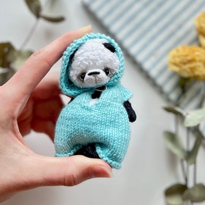 Puede incluir: Un pequeño peluche de panda blanco y negro con un suéter de punto azul claro con capucha.