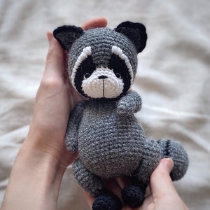 May include: A gray crocheted raccoon plush toy with black eyes, a white nose, and black ears. The toy is being held by two hands.